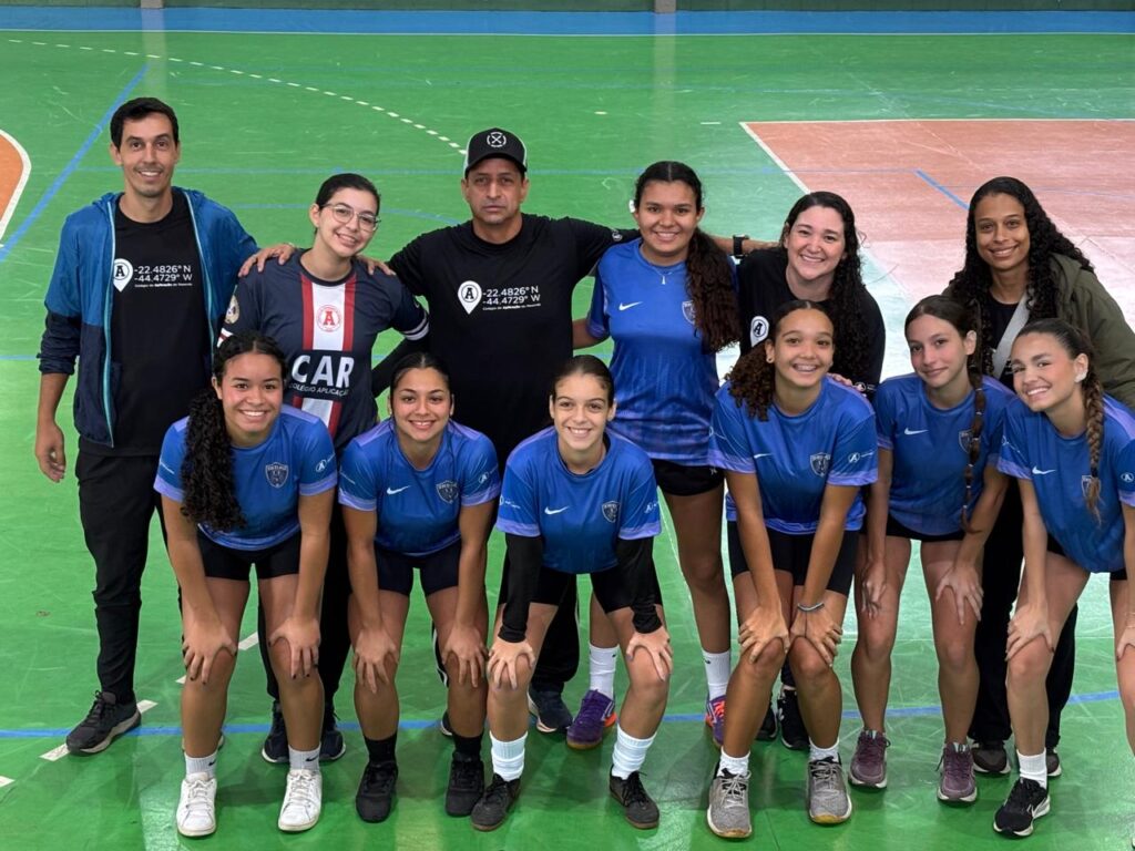 Equipe feminina de atletas escolares do Colégio de Aplicação de Resende posando para foto junto com seus treinadores e comissão técnica em uma quadra de esportes verde durante o JEPAR.
