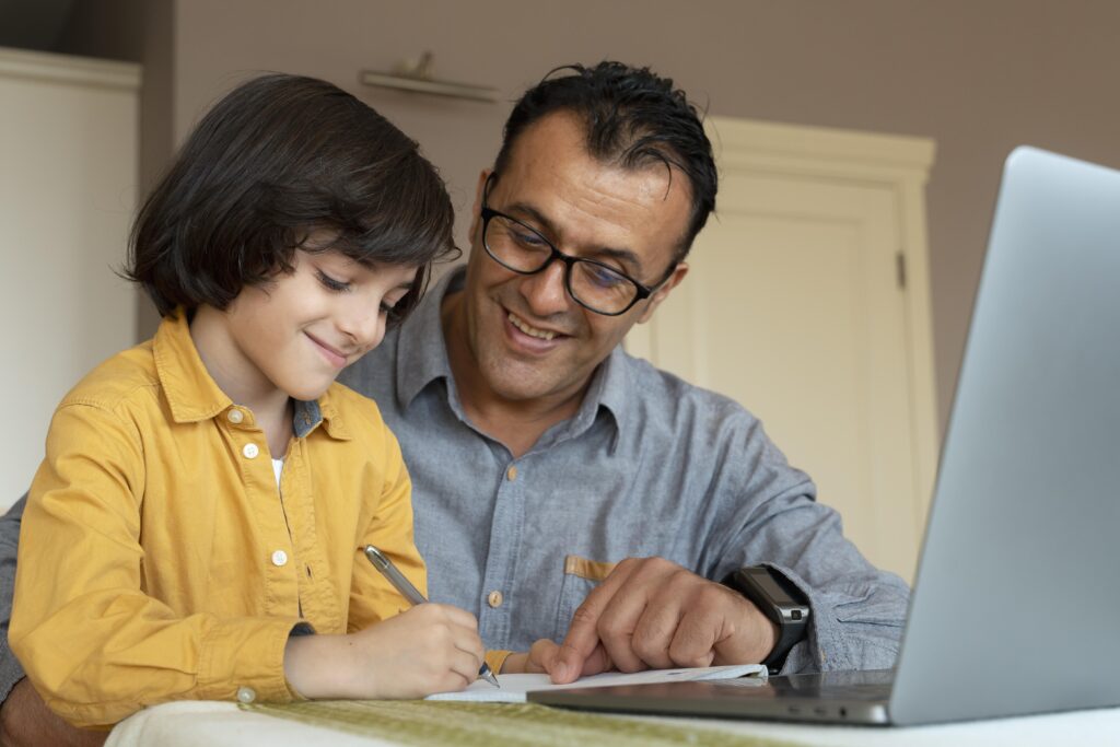Pai sorrindo ao lado do filho que escreve no caderno, com um notebook aberto na mesa, mostrando apoio familiar e o uso de tecnologia na rotina de estudos para crianças.
