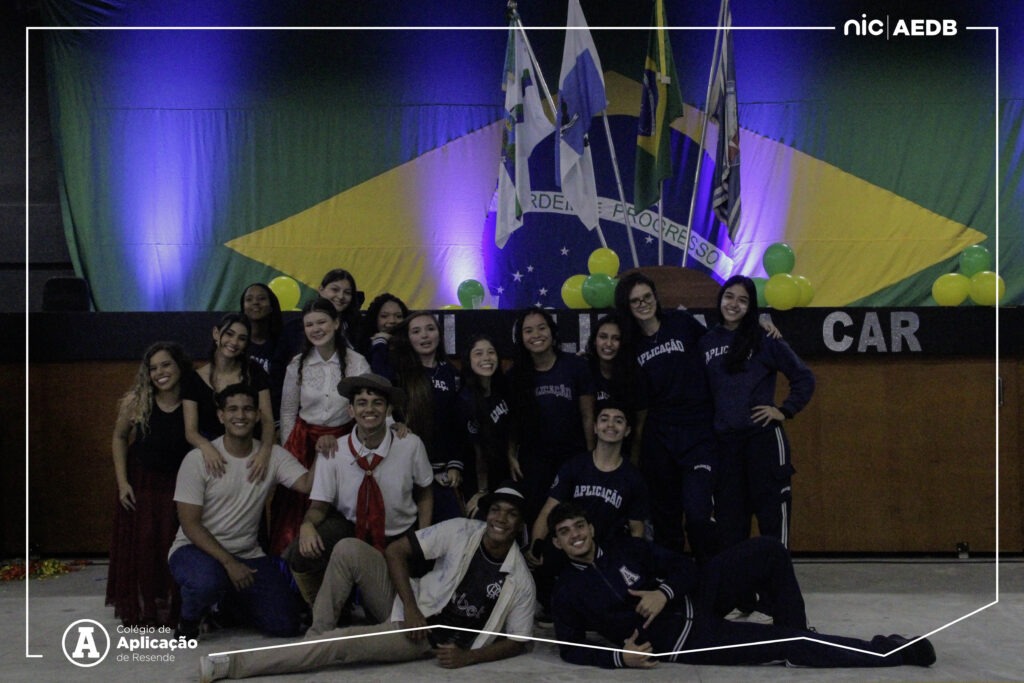 Grupo de alunos do Ensino Medio posando sorridentes em frente ao palco com a bandeira do Brasil na abertura das Olimpiadas Escolares do Colegio de Aplicacao de Resende.