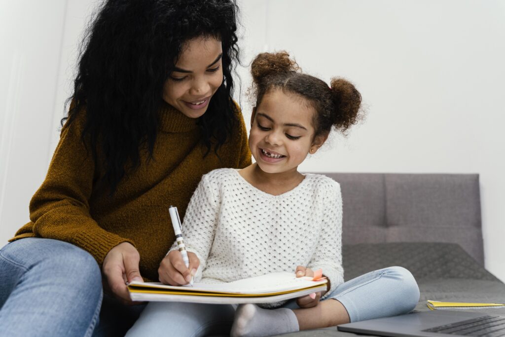 Mãe ou irmã mais velha sorrindo e ajudando uma criança pequena a escrever no caderno durante o estudo em casa, destacando a parceria entre família e escola.