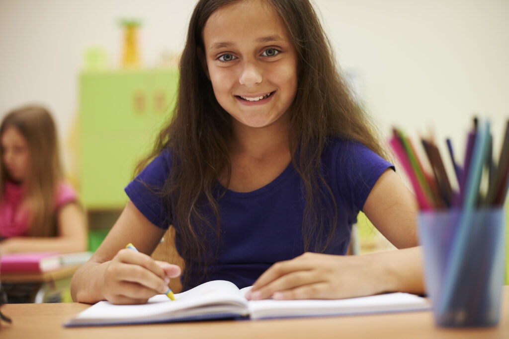 Menina sorrindo com orgulho e caneta na mão, olhando para o caderno, demonstrando motivação com os hábitos de estudo em sala de aula do ensino fundamental.