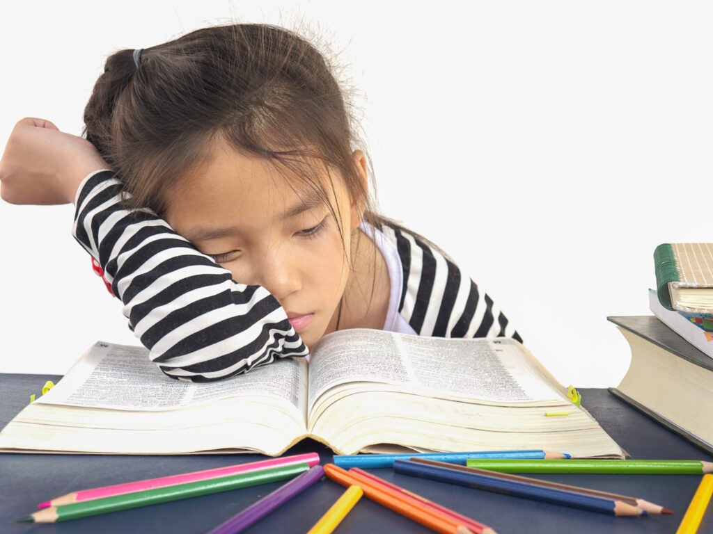 Menina cansada dormindo em cima de um livro aberto com lápis de cor ao redor, ilustrando a falta de uma rotina de estudos para crianças organizada.