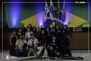 Grupo de alunos do Ensino Medio posando sorridentes em frente ao palco com a bandeira do Brasil na abertura das Olimpiadas Escolares do Colegio de Aplicacao de Resende.