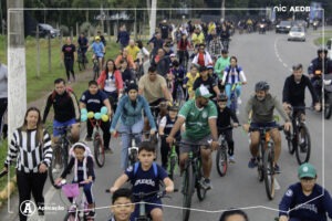 Grande grupo de estudantes de várias idades, familiares e educadores do Colégio de Aplicação de Resende pedalando juntos em uma avenida durante o tradicional Passeio Ciclístico da comunidade escolar.